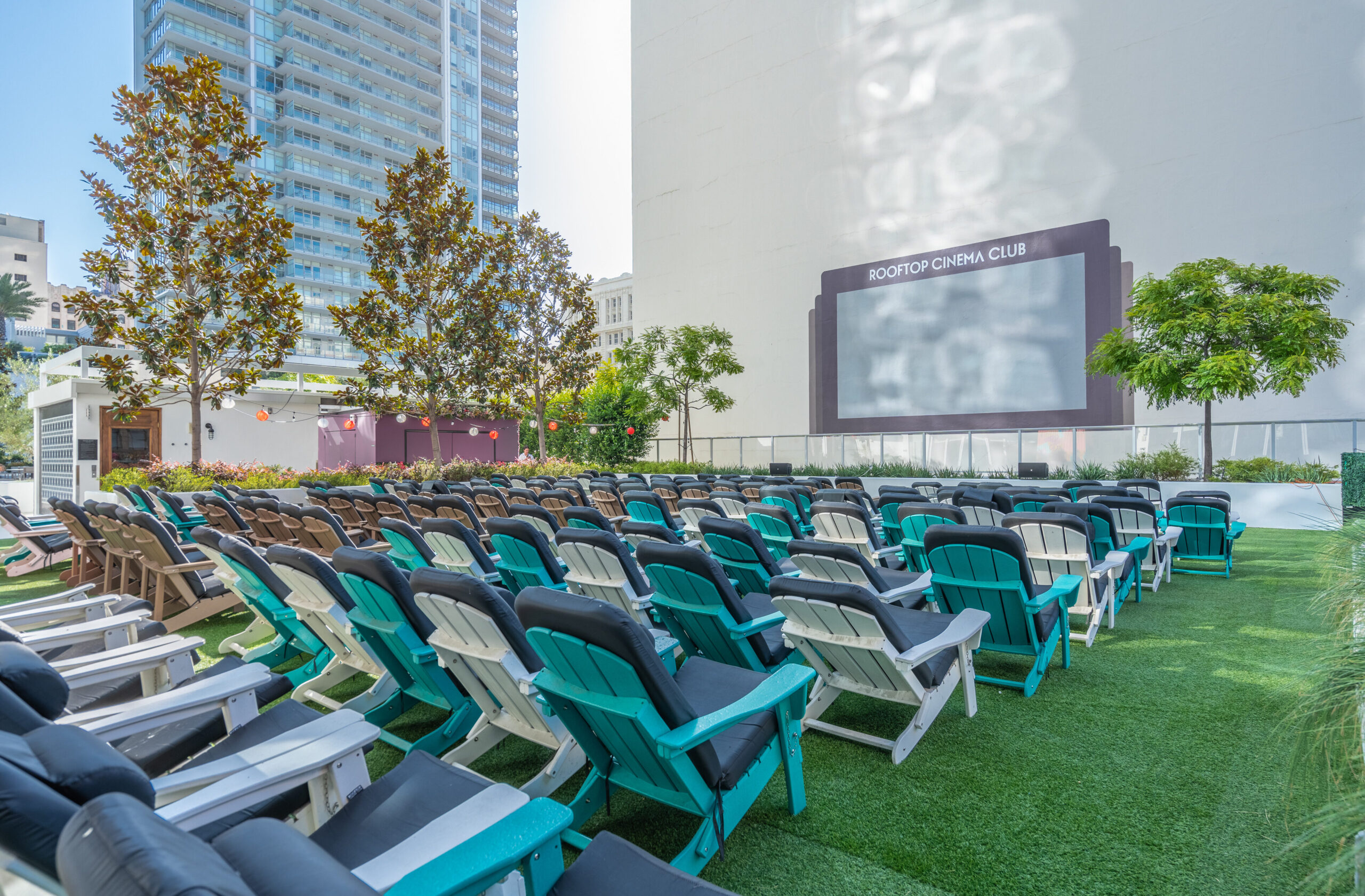 Rows of colorful cushioned chairs face a large outdoor movie screen at a rooftop cinema, surrounded by greenery and city buildings in the background under clear daylight.