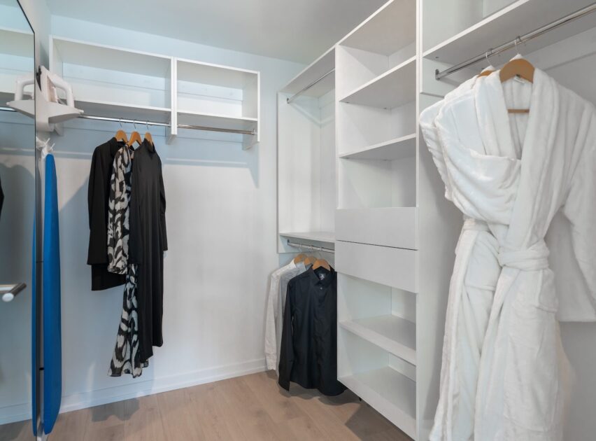 Modern walk-in closet with open shelves, hanging rods, and a few clothes, including black and patterned garments. A white bathrobe hangs on a hook, and an ironing board is visible on the left. The floor is light wood.