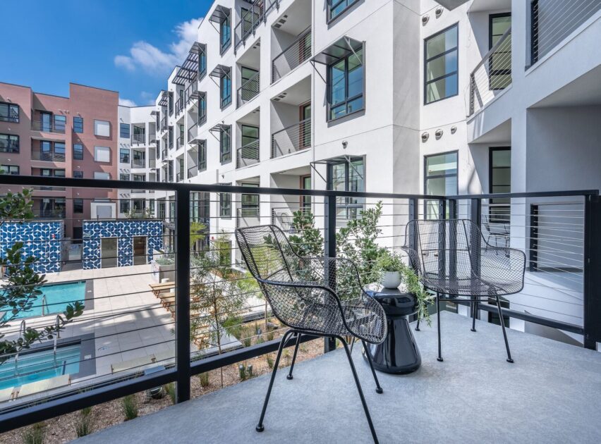 Modern apartment balcony with two black metal chairs and a small round table, overlooking a courtyard with a swimming pool, surrounded by white multi-story buildings under a blue sky.