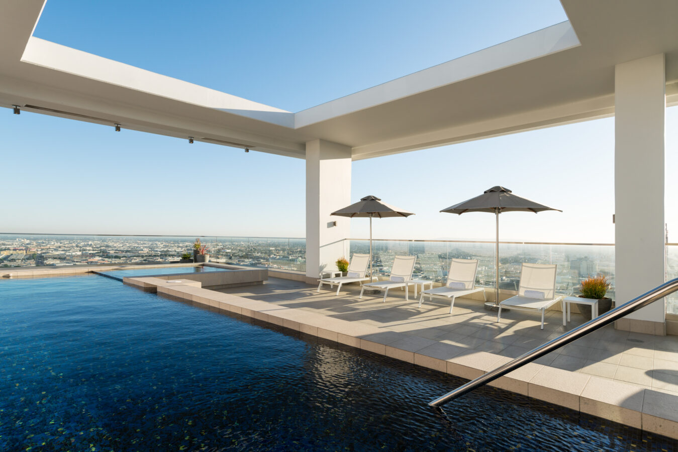 Rooftop infinity pool with lounge chairs and umbrellas under a modern white pergola, overlooking a cityscape on a clear, sunny day.