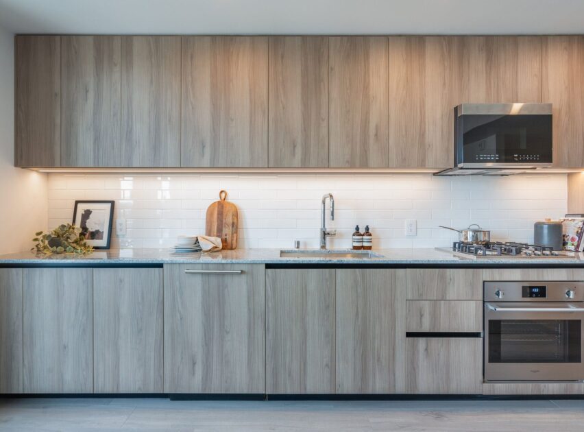 Modern kitchen with light wood cabinets, white subway tile backsplash, built-in oven, gas stove, sink, and minimal decor including a cutting board, framed picture, and a small plant on the countertop.