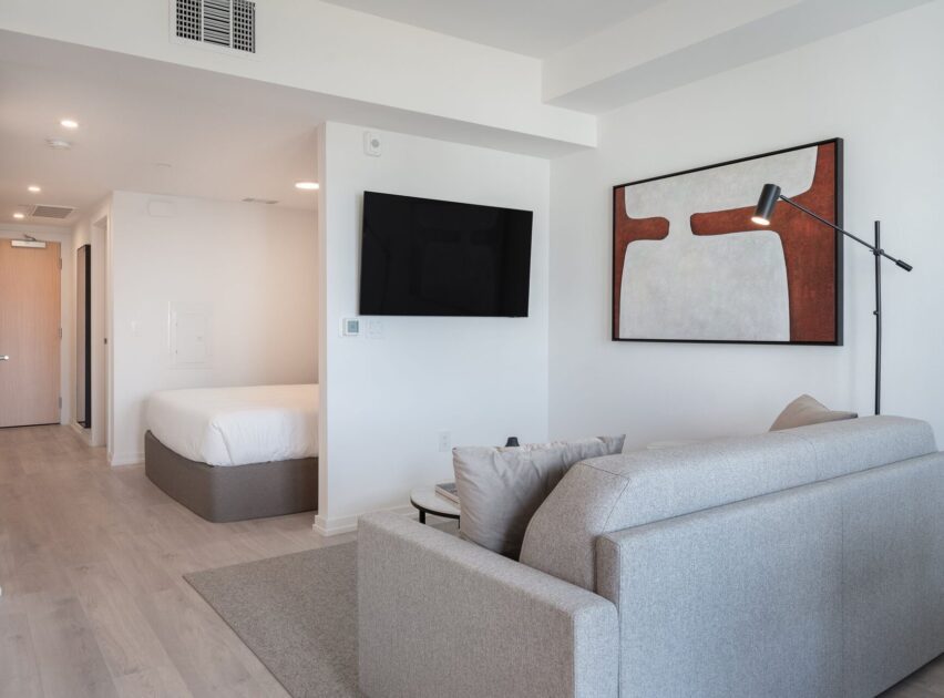A modern studio apartment with light wood flooring, a bed in an alcove, a wall-mounted TV, a gray sofa, a floor lamp, and a large abstract painting with white and rust tones above the couch.