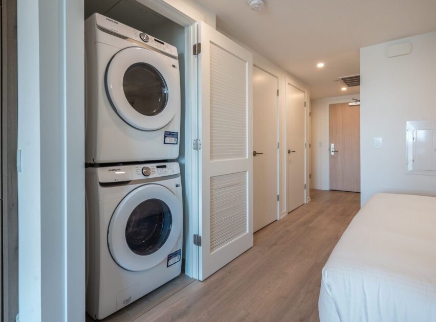 A modern laundry area in an apartment features a stacked washer and dryer next to white doors and a hallway with light wood flooring, visible from the edge of a bedroom.