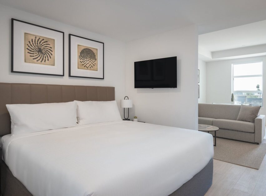 A modern hotel room with a large neatly made bed, two framed abstract artworks above the headboard, a wall-mounted TV, a bedside lamp, and a sitting area with a sofa and coffee table near a window.