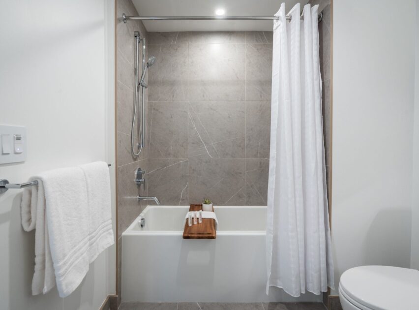 Modern bathroom with a bathtub, gray marble tile wall, white shower curtain, towel on a rack, and a wooden bath tray holding towels and a small potted plant. Toilet partially visible on the right.