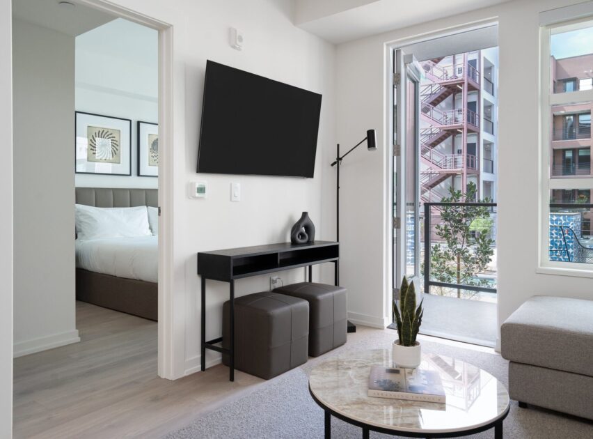 Modern living room with a wall-mounted TV, black console table, two stools, and a round coffee table with a plant. An open door leads to a bedroom, and sliding glass doors open to a balcony with city views.