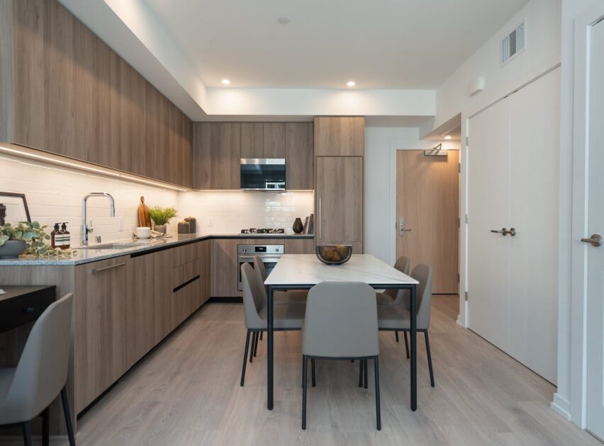 Modern kitchen and dining area with light wood cabinets, white countertops, integrated appliances, and a rectangular dining table with six gray chairs. Bright lighting and minimalist decor create a clean, contemporary look.