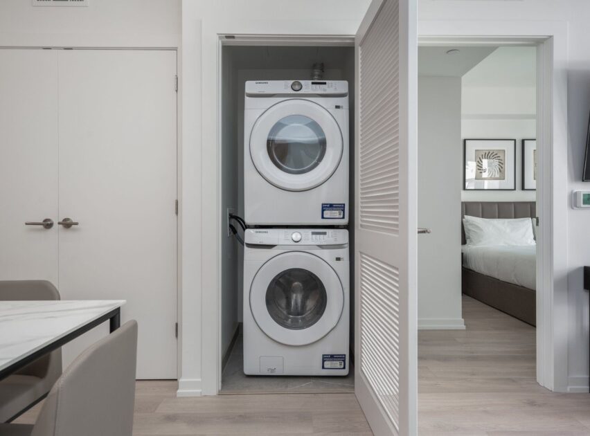 A stacked washer and dryer are set in a closet with a partially open louvered door, next to a bedroom and a dining area in a modern apartment.