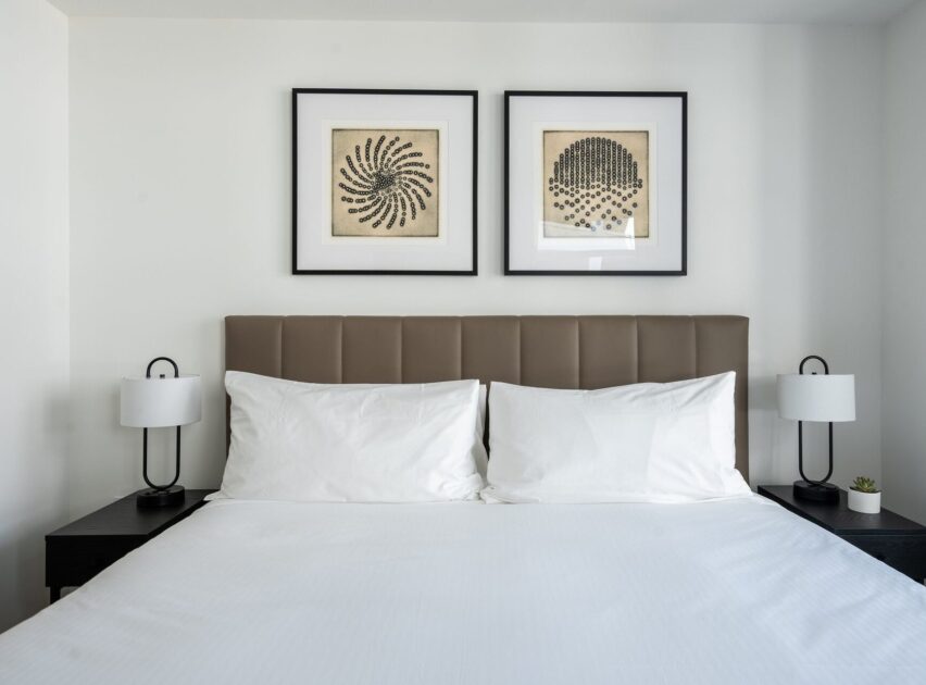 A neatly made bed with white linens and two pillows, flanked by black nightstands with modern white lamps. Two framed abstract artworks hang on the white wall above the headboard.