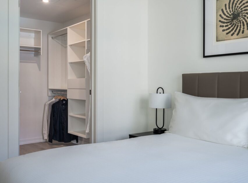A neatly made bed with white linens beside a black nightstand and lamp; in the background, there is an open walk-in closet with hanging clothes and shelving. A framed artwork hangs on the wall above the bed.