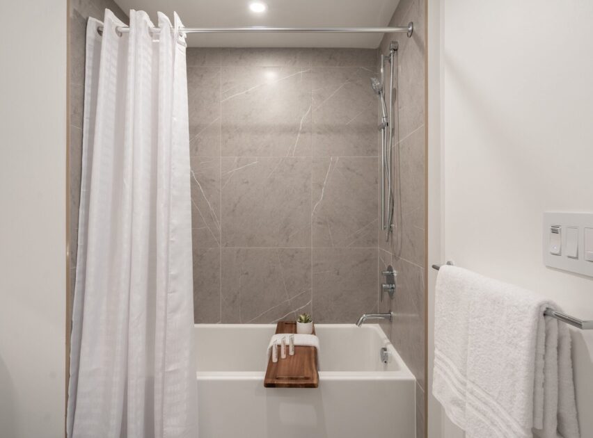 Modern bathroom with gray tile walls, a white bathtub, white shower curtain, towel rack with a white towel, and a wooden bath tray holding a folded towel, gloves, and a small plant.