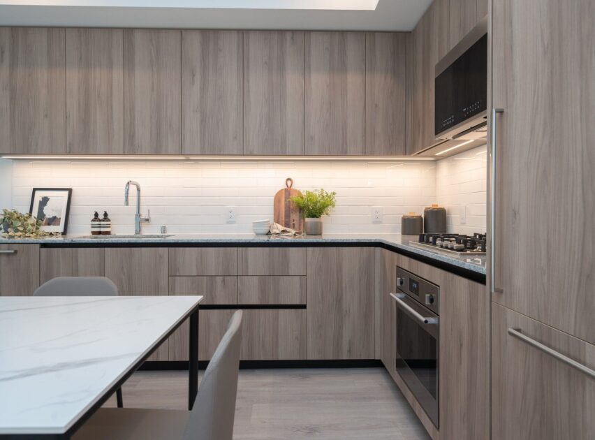 Modern kitchen with light wood cabinets, white subway tile backsplash, built-in oven and stove, marble dining table, minimalist decor, and under-cabinet lighting. A plant and kitchen accessories are arranged on the counter.