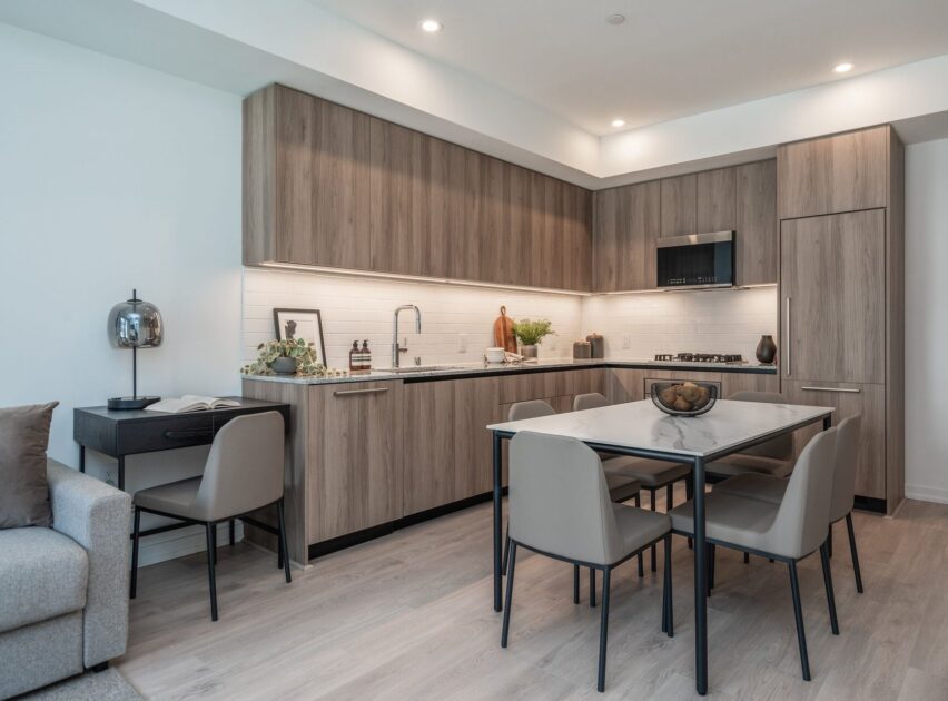 Modern kitchen with light wood cabinets, white backsplash, and built-in appliances. A dining table with four beige chairs sits in the center. A small desk with a lamp is on the left near a gray sofa. The space is bright and tidy.