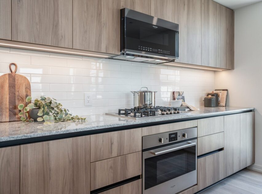 Modern kitchen with light wood cabinets, a built-in oven, a gas cooktop with a pot, a black range hood, white subway tile backsplash, and decorative items like a plant and cutting boards on the countertop.