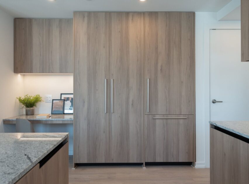 Contemporary kitchen with light wood cabinets, sleek metal handles, granite countertops, and a small built-in desk area with a plant and framed photos under soft lighting.