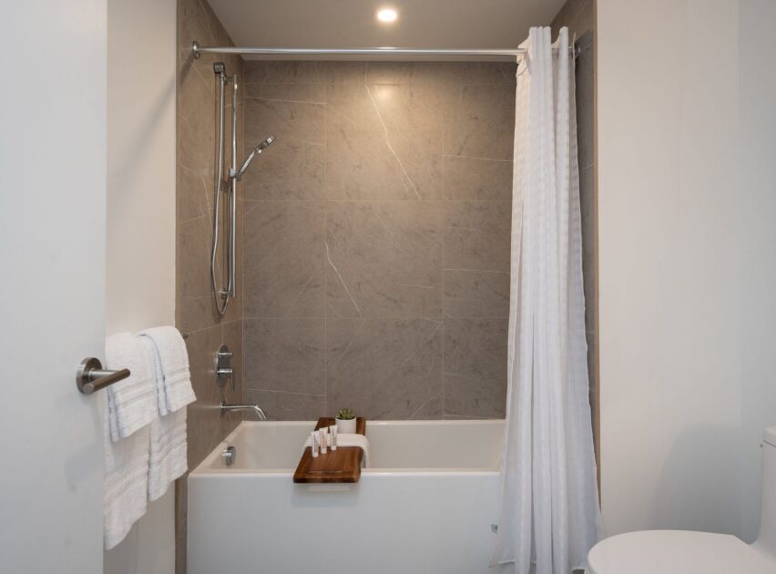 Modern bathroom with a bathtub, gray tiled wall, mounted showerhead, white shower curtain, towel on a rack, and a wooden bath tray holding toiletries and a small plant.