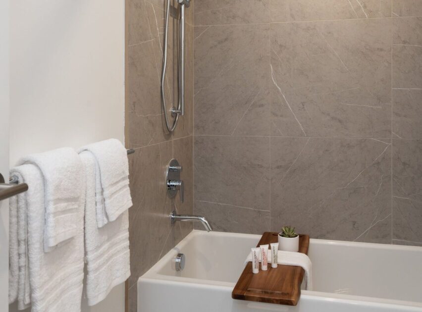 Modern bathroom with a gray tiled shower, white towels hanging on a rack, and a white tub. A wooden bath tray holds small toiletries and a small potted succulent.