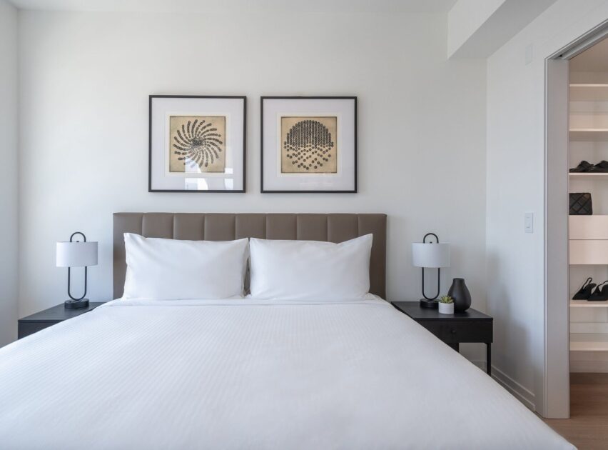 A modern, minimalistic bedroom with a neatly made bed, white linens, two black bedside tables with lamps, and two framed abstract artworks above the headboard. An open closet is visible to the right.