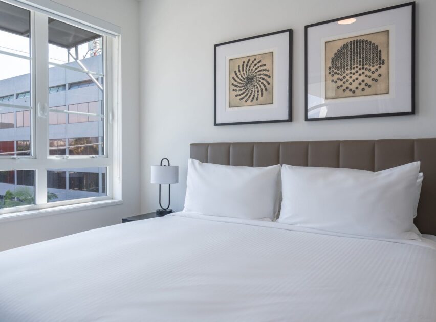 A neatly made bed with white linens and two pillows sits next to a window. Two framed abstract artworks hang above the padded headboard, and a small lamp is on the bedside table. An office building is visible outside.