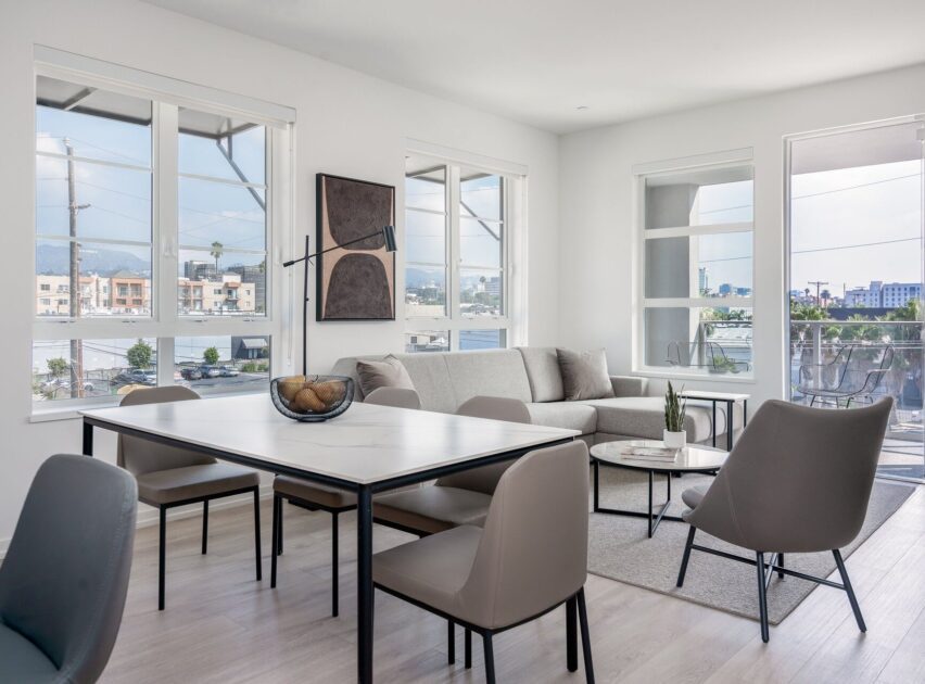 Bright, modern living and dining area with large windows, light wood floors, a gray sofa, minimalist decor, dining table with chairs, and a small balcony visible outside.