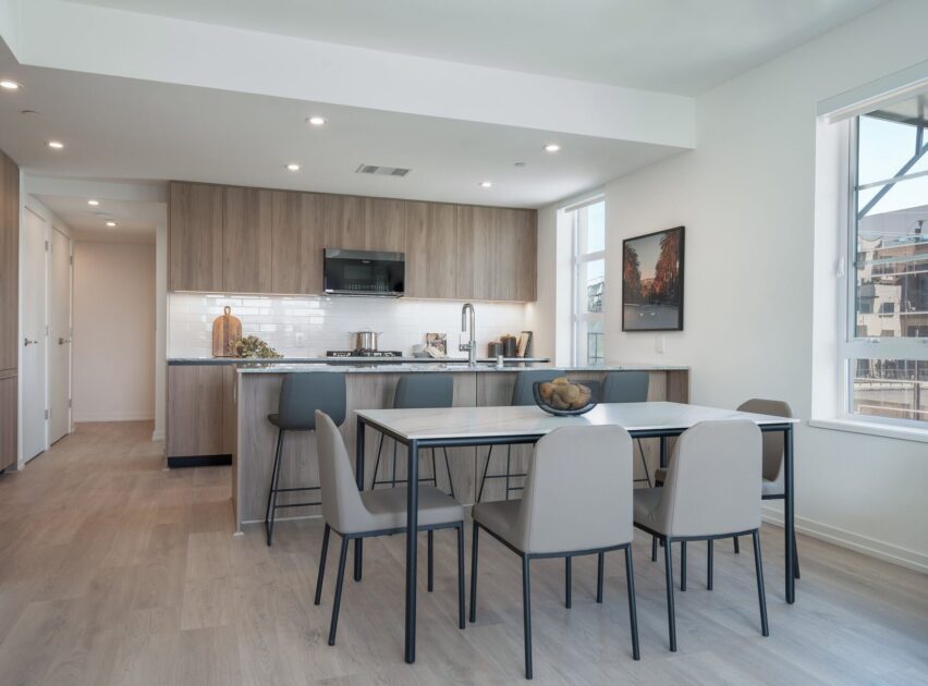 Modern kitchen with light wood cabinets, white countertops, and stainless steel appliances. A dining table with six gray chairs is in the foreground. Large windows provide natural light, and decor is minimal and contemporary.