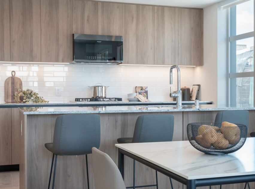 Modern kitchen with light wood cabinets, marble island countertop, and gray bar stools. A basket of lemons is on the dining table, and sunlight streams in through large windows. Stainless steel appliances are visible.