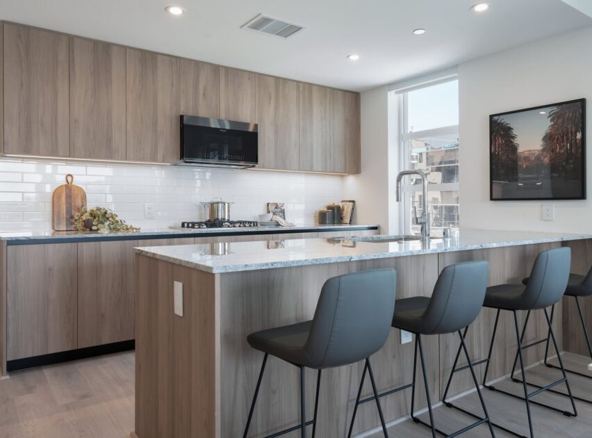 Modern kitchen with light wood cabinets, white countertops, a large island with gray bar stools, stainless steel appliances, and a window letting in natural light. A framed picture and decorative items are on display.