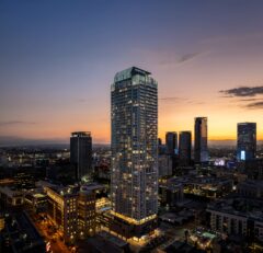 A tall, modern skyscraper illuminated at dusk towers over a cityscape with other high-rise buildings, glowing lights, and a colorful sunset sky in the background.
