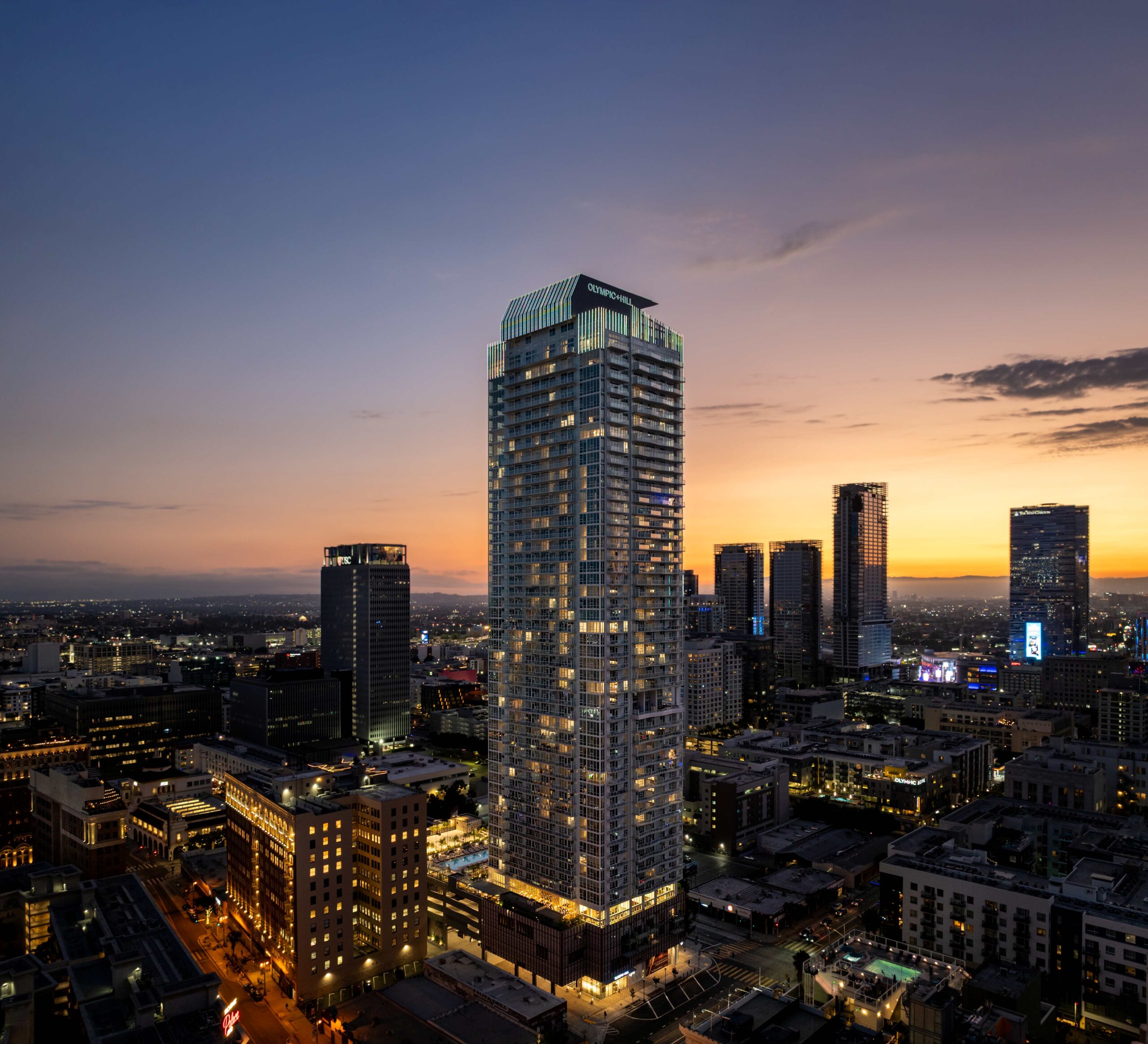 A tall, modern skyscraper illuminated at dusk towers over a cityscape with other high-rise buildings, glowing lights, and a colorful sunset sky in the background.