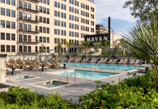 A rooftop pool area with lounge chairs and umbrellas surrounded by greenery, set against a large beige building. A sign reading Mayan Theater is visible in the background.