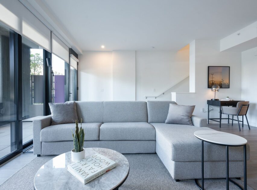 A modern living room with a light gray sectional sofa, round marble coffee tables, a small potted plant, and books. Floor-to-ceiling windows provide natural light, and a desk and chair are in the background near a staircase.