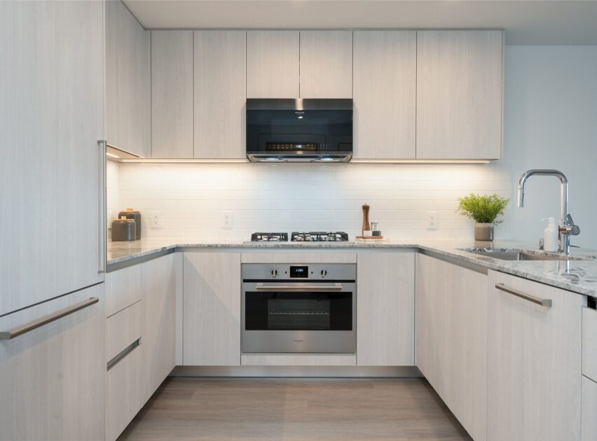 Modern kitchen with light wood cabinets, built-in oven, gas stovetop, stainless steel appliances, and marble countertops. Under-cabinet lighting illuminates a white subway tile backsplash. A plant and kitchen accessories are on the counter.