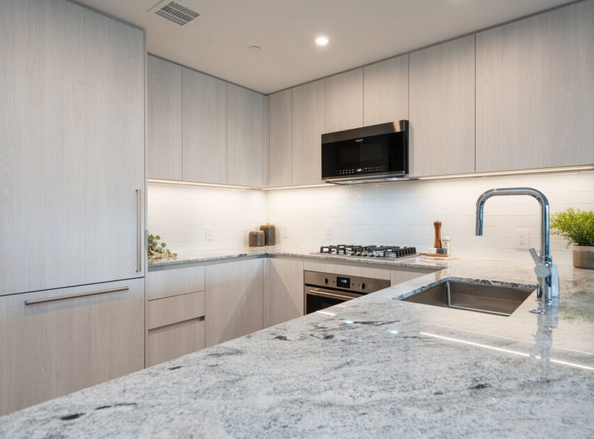 Modern kitchen with light wood cabinets, marble countertops, stainless steel appliances, a built-in oven, gas stove, microwave, under-cabinet lighting, and a sink with a sleek faucet; small potted plant on the counter.