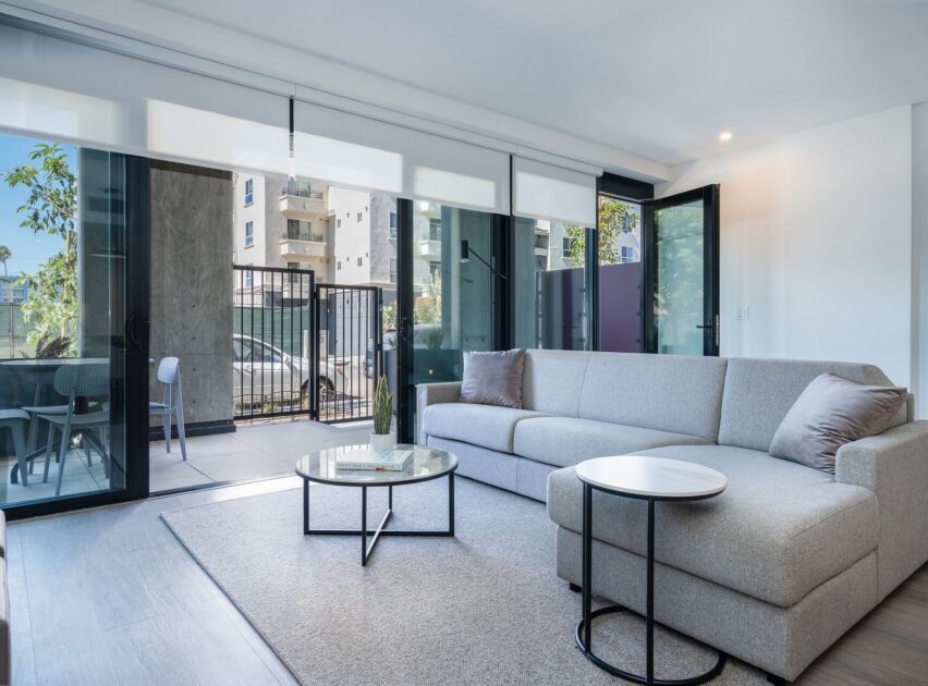 Modern living room with a light gray sectional sofa, round coffee table, and side table, featuring large sliding glass doors that open to a private patio with outdoor seating. Natural light fills the space.