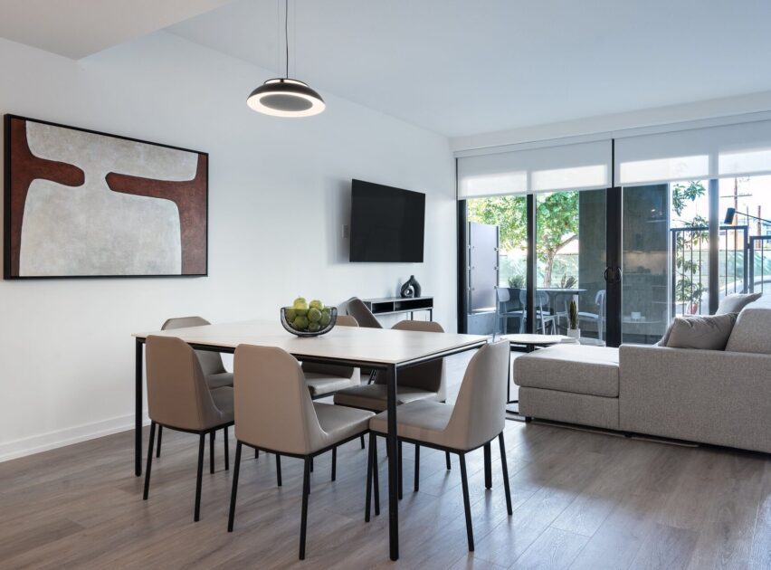 Modern dining and living area with a table and six beige chairs, a bowl of green apples, wall-mounted TV, abstract art, large windows with blinds, and a light gray sectional sofa facing a patio.