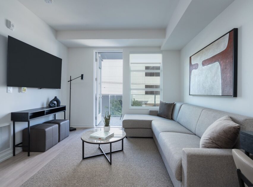 A modern living room with a gray sectional sofa, abstract wall art, a round coffee table, a TV mounted on the wall, and large windows letting in natural light.