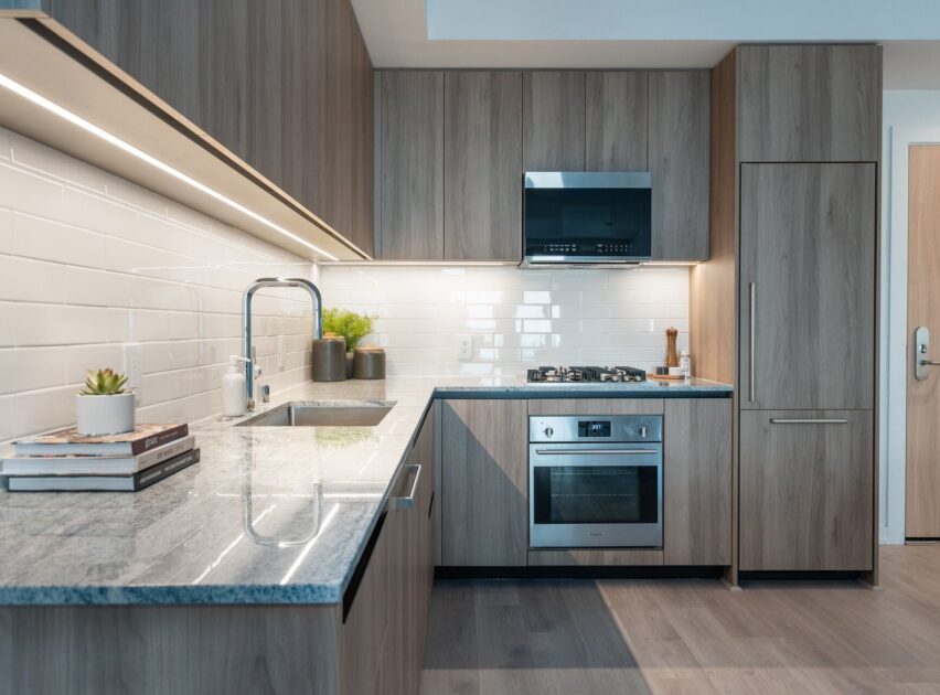 Modern kitchen with light wood cabinets, white subway tile backsplash, built-in appliances, and a gray countertop. There are a few books, a small plant, and a cooktop with an oven below on the right side.