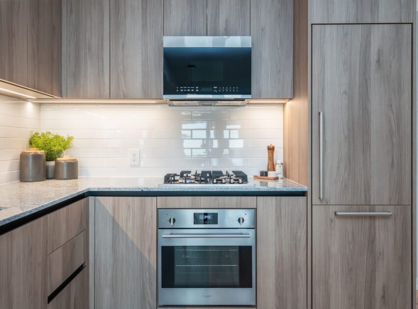 Modern kitchen with light wood cabinets, a built-in oven, gas stovetop, and stainless steel range hood. White subway tile backsplash, countertop with a potted plant, utensils, and integrated refrigerator are also visible.