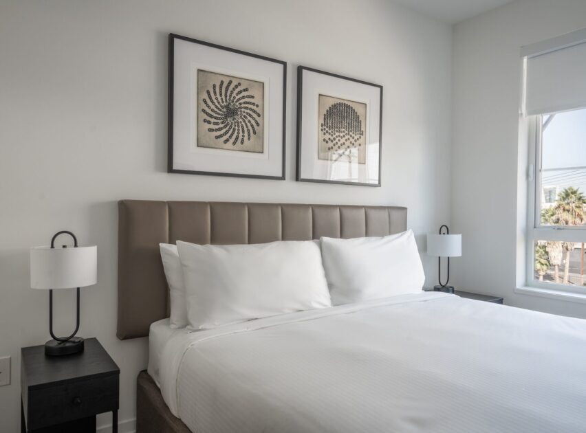 A modern, minimal bedroom with a neatly made bed, white linens, padded headboard, two black nightstands with lamps, and two abstract framed artworks above the bed. Sunlight streams in from a window on the right.