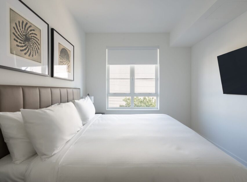 A modern, minimal bedroom with a neatly made bed, white bedding, two framed artworks above the headboard, a window with a shade, and a wall-mounted TV on the right.
