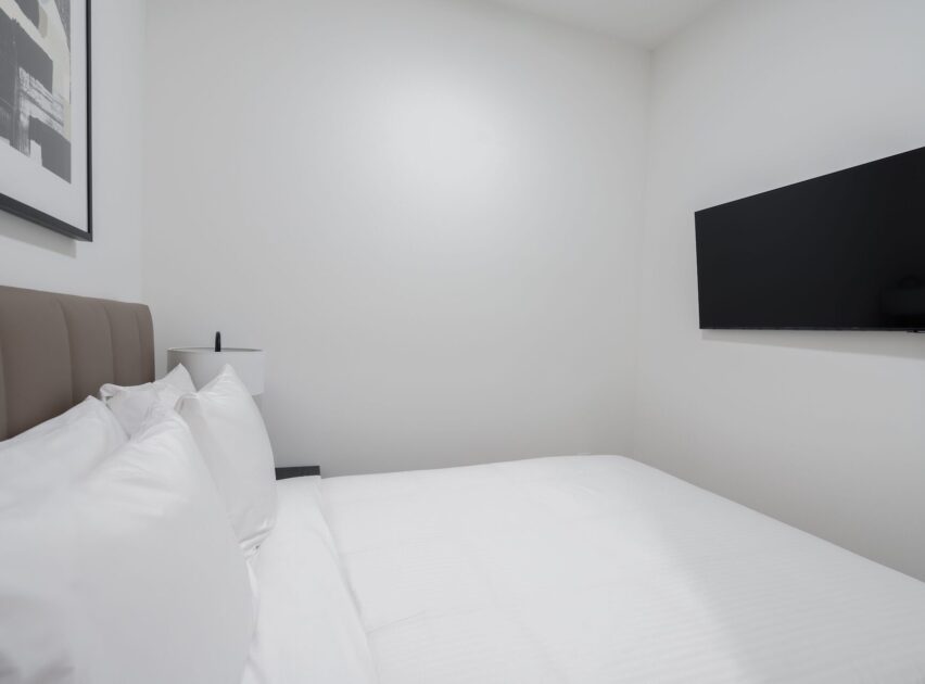 A minimal, modern bedroom with a large bed covered in white linens, a brown padded headboard, a wall-mounted TV, a lamp on a nightstand, and a framed abstract artwork on a white wall.