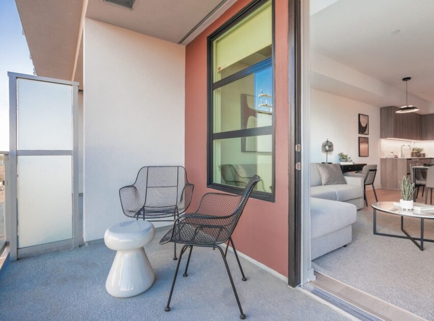 A modern apartment balcony with two black wire chairs, a small white side table, and frosted glass panel. The sliding door leads to a living room with a gray sofa and open kitchen.