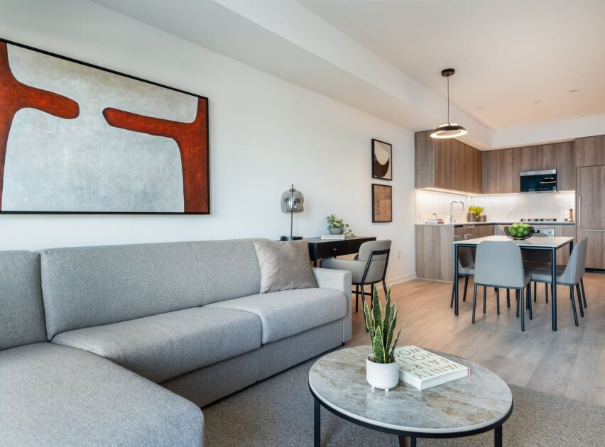 Modern living room with a light gray sectional sofa, round coffee table with a plant and book, abstract wall art, and open kitchen with wooden cabinets and dining table in the background.