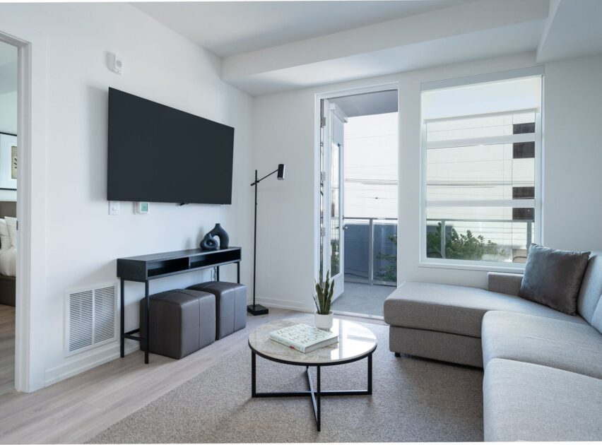 Modern living room with a wall-mounted TV, a sectional sofa, a round coffee table with a plant, and large windows letting in natural light. A bedroom is visible through an open door to the left.
