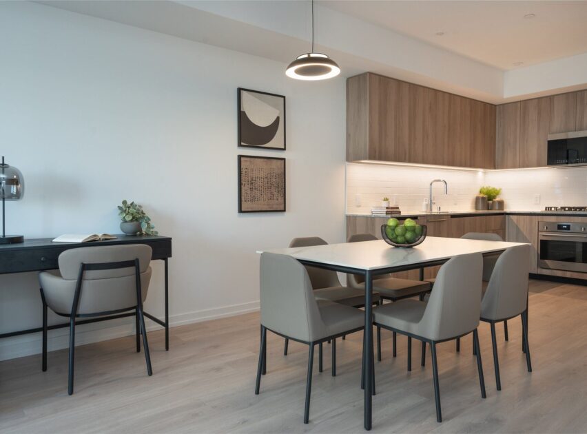 Modern kitchen and dining area with light wood cabinets, a black-edged dining table with six beige chairs, a bowl of green apples as centerpiece, and a small desk setup with a chair and lamp on the left.