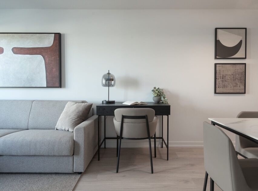 Minimalist living space with a light gray sofa, a black desk with a lamp, a chair, and a plant. Abstract art hangs on the white wall above the sofa and desk, with a dining table partially visible on the right.
