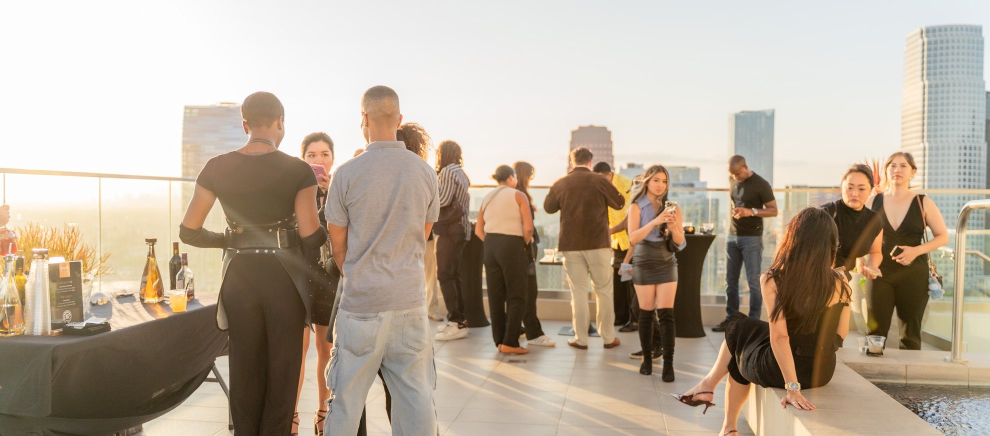A group of people dressed in semi-formal attire socialize on a rooftop terrace at sunset, with city buildings in the background and tables set with drinks and food on the side.