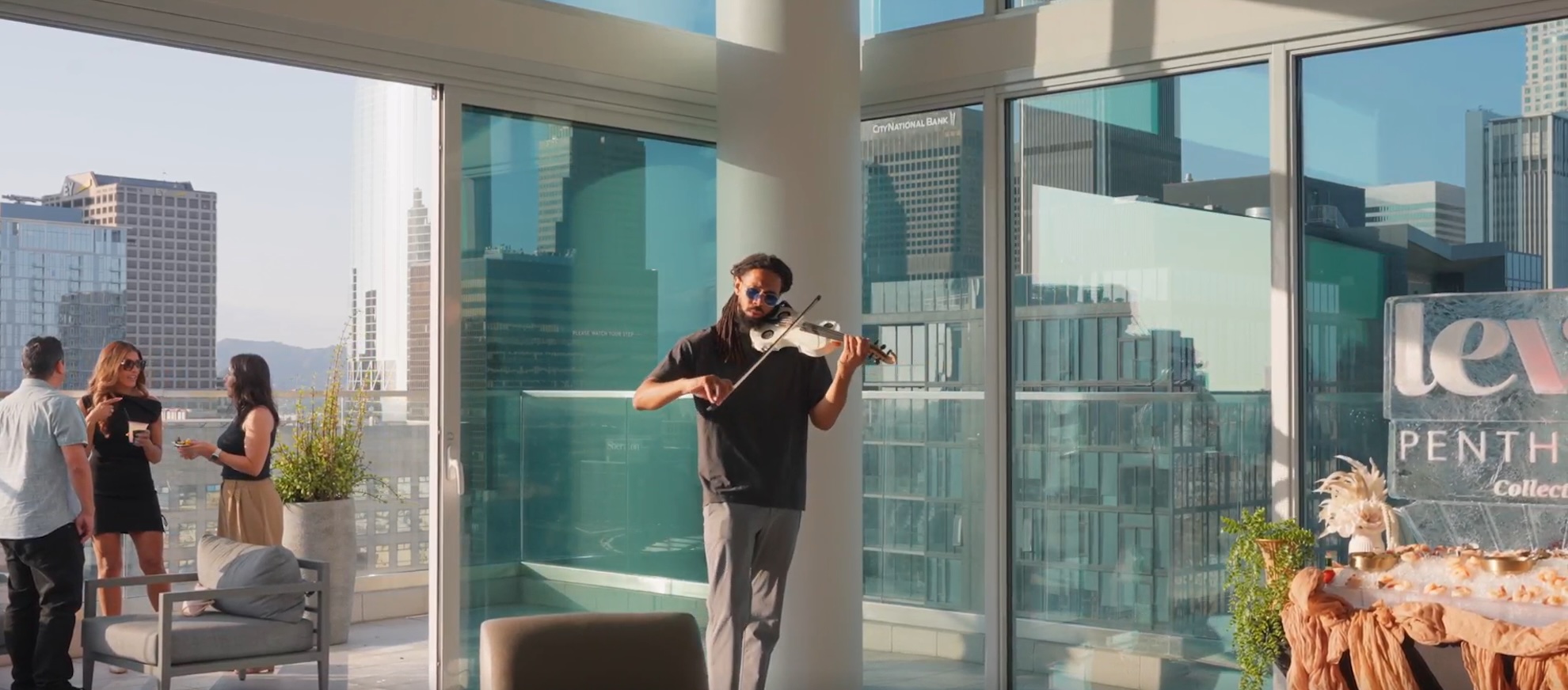 A man plays a violin inside a modern apartment with large glass windows, overlooking a city skyline. On the balcony outside, three people socialize, enjoying the sunlight and urban view.