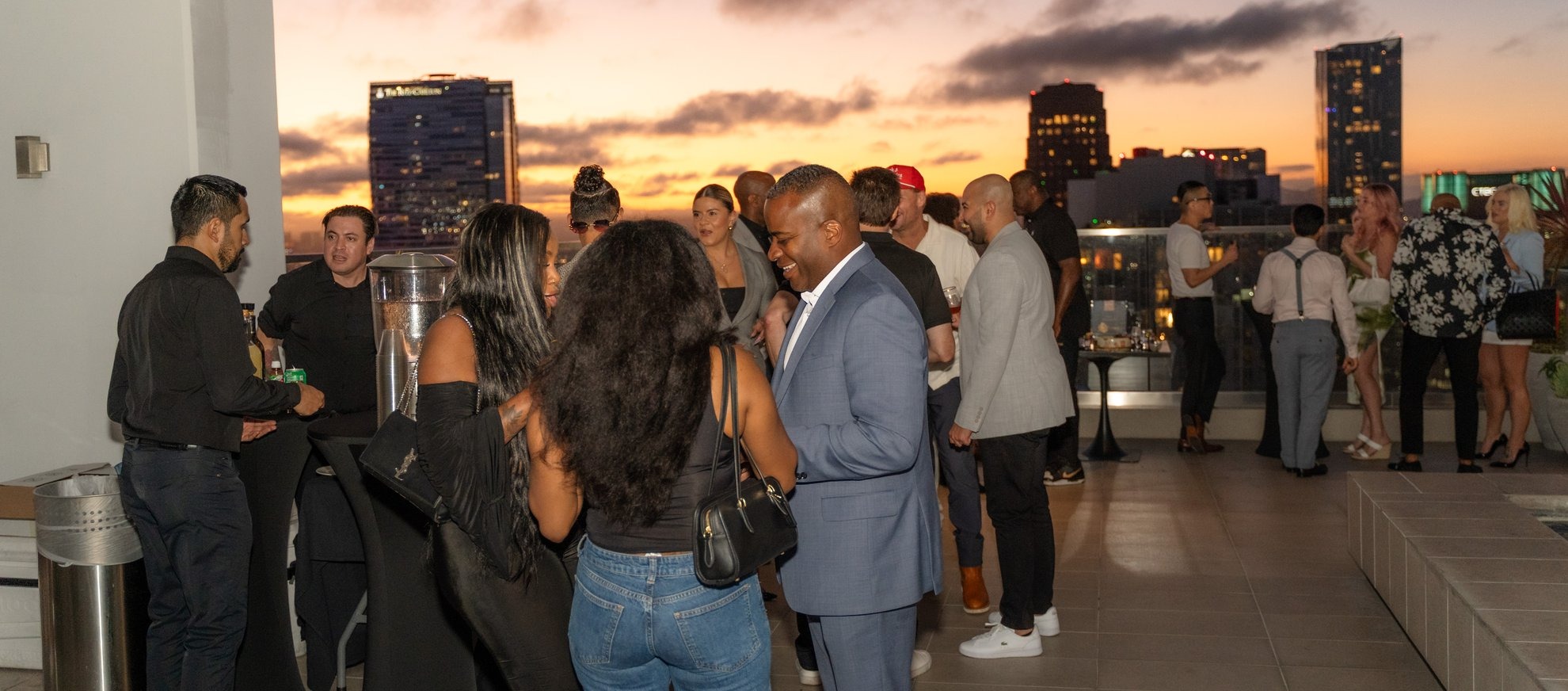 A group of people socialize on a rooftop at sunset, with city buildings and a colorful sky in the background. Some are chatting in small groups, while others stand by the railing enjoying the view.