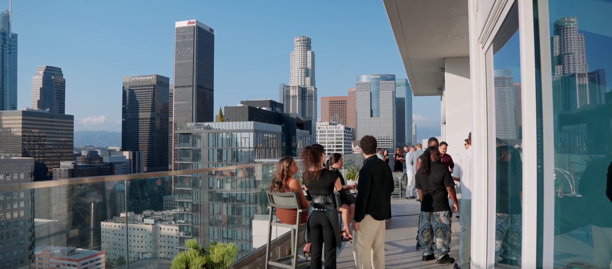 People socialize on a modern rooftop terrace overlooking a city skyline with tall glass and concrete buildings under a clear blue sky.